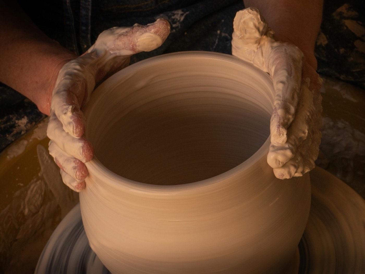 A pair of clay-covered hands steadying a pot as it turns on a potter's wheel